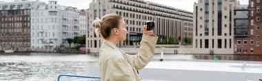 Trençkotlu bir kadının Berlin 'de cep telefonuyla fotoğraf çekmesi, afiş. 