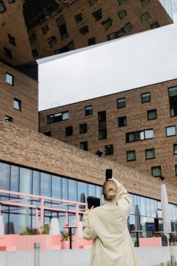 Trençkotlu genç turistin Berlin 'deki binanın yakınındaki akıllı telefondan fotoğraf çekerken görüntüsü. 