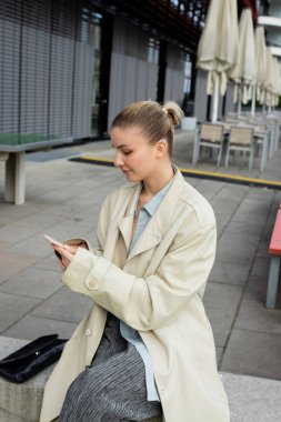 Berlin 'de debriyaj çantasının yanında cep telefonu tutan trençkotlu genç bir kadın. 