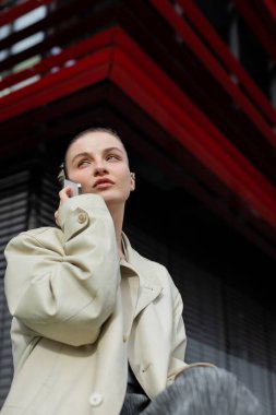 Trençkotlu şık bir kadının açık havada akıllı telefondan konuşmasının düşük açılı görüntüsü. 