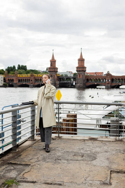 Young woman in trench coat talking on smartphone on pier in Berlin 
