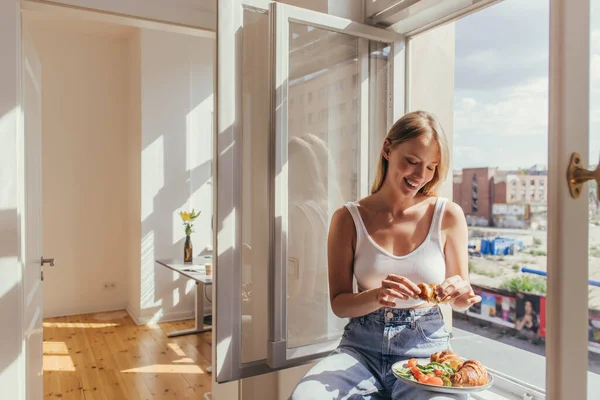 Leende Kvinna Håller Croissant När Hon Sitter Fönsterbrädan Hemma — Stockfoto