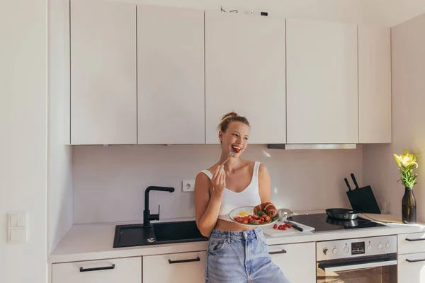 笑顔ブロンド女性食べるチェリートマトとホールディングプレートともに卵とクロワッサンでキッチン — ストック写真