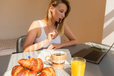 Diz üstü bilgisayarın yanında elinde kalem ve lezzetli bir kahvaltı tutan sarışın kadın. 