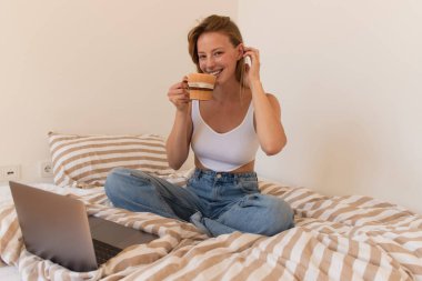 Yatakta dizüstü bilgisayarın yanında kahve içerken kameraya gülümseyen genç serbest yazar. 