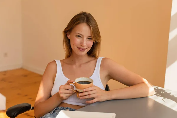 金发女人顶着一杯咖啡 看着家里笔记本旁边的相机 — 图库照片
