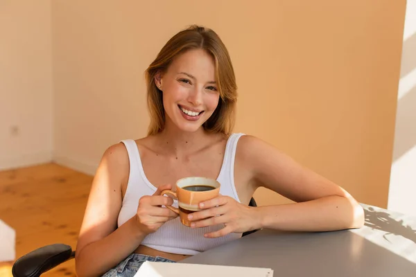 Vrolijke Blonde Vrouw Met Kopje Koffie Buurt Notebook Tafel Thuis — Stockfoto
