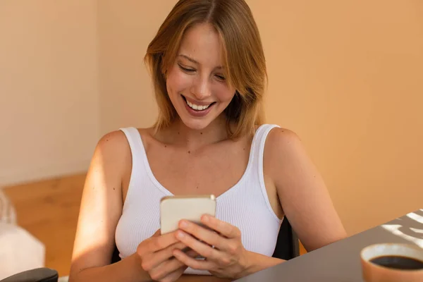 Gülümseyen Sarışın Kadın Evde Bulanık Kahvenin Yanında Akıllı Telefon Kullanıyor — Stok fotoğraf