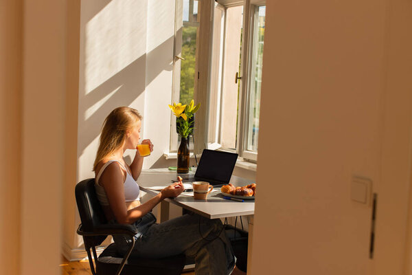 Вид сбоку на молодого фрилансера, пьющего апельсиновый сок рядом с устройствами и круассанами дома 