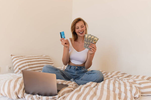 Веселая женщина в топе и джинсах с кредиткой и долларовыми банкнотами рядом с ноутбуком на кровати 