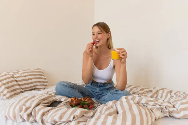 Smiling woman holding cherry tomato and orange juice near croissants and smartphone on bed