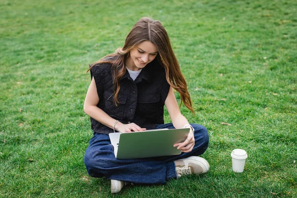 Feliz joven mujer usando el ordenador portátil mientras está sentado en el césped - foto de stock