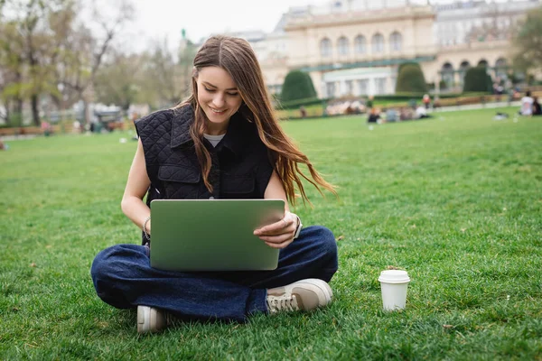 Весела молода жінка використовує ноутбук, сидячи на газоні — стокове фото