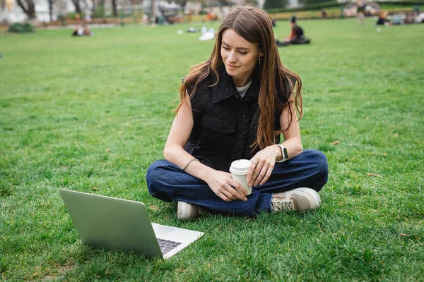 Cheerful young woman holding paper cup and looking at laptop while sitting on lawn — Stock Photo