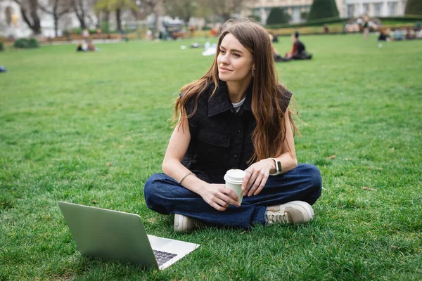 Веселая молодая женщина держит бумажную чашку возле ноутбука, сидя на газоне — стоковое фото
