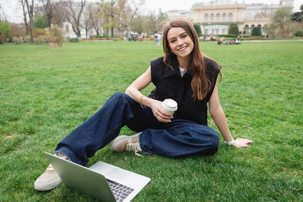 Sonriente mujer joven sosteniendo taza de papel cerca de la computadora portátil mientras está sentado en el césped - foto de stock