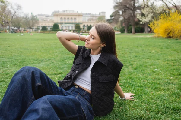 Glückliche junge Frau in ärmelloser Jacke auf Rasen sitzend — Stockfoto