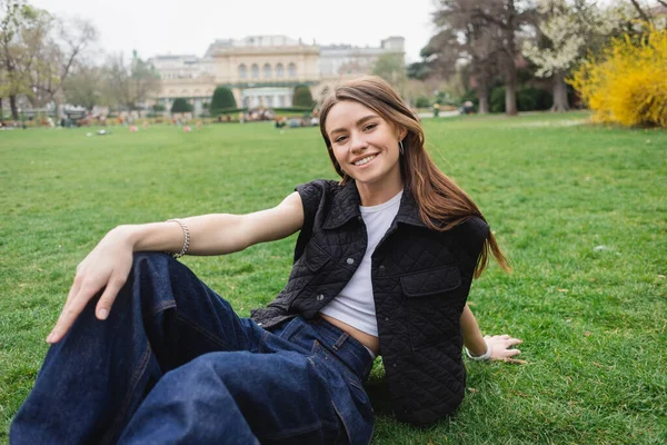 Радостная молодая женщина в куртке без рукавов сидит на газоне — стоковое фото