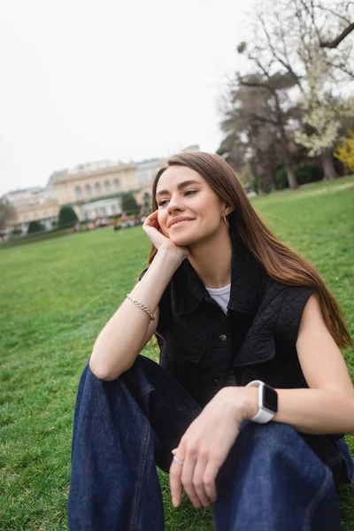 Довольная молодая женщина в куртке без рукавов сидит на газоне — стоковое фото