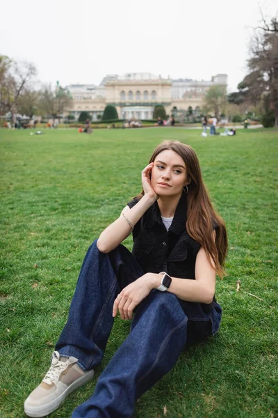 Thoughtful young woman in sleeveless jacket sitting on lawn — Stock Photo