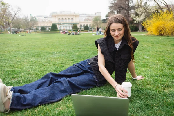 Sorridente giovane donna in giacca senza maniche che tiene tazza di carta e poggia sul prato vicino al computer portatile — Foto stock