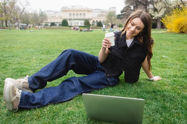 Feliz joven mujer en chaqueta sin mangas sosteniendo taza de papel y descansando en el césped cerca de la computadora portátil - foto de stock