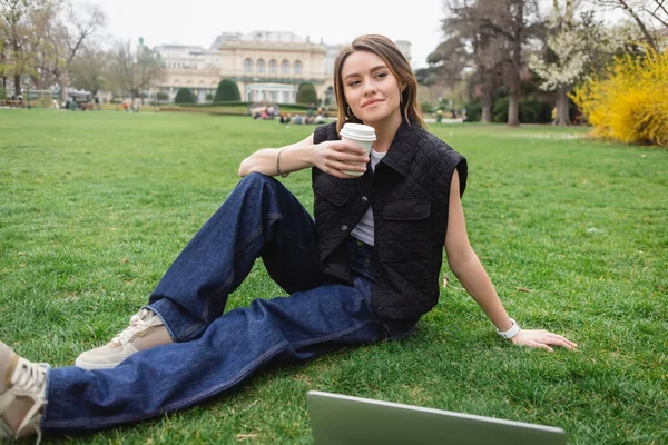 Jeune femme réfléchie dans une veste sans manches tenant tasse en papier et reposant sur la pelouse près d'un ordinateur portable — Photo de stock