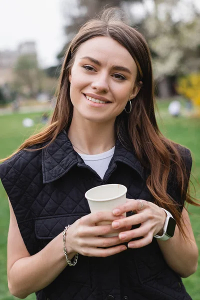 Young cheerful woman in sleeveless jacket holding coffee to go in green park — Stock Photo