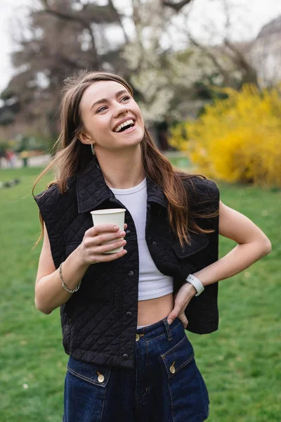 Молодая радостная женщина в куртке без рукавов держит кофе, чтобы пойти в зеленый парк — стоковое фото