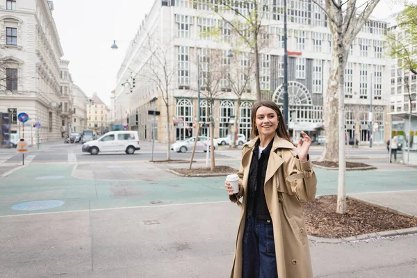Mujer joven feliz en gabardina elegante caminando con café para ir y saludando de la mano en la ciudad europea - foto de stock