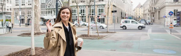 Mujer joven feliz con elegante gabardina sosteniendo café para ir y saludando de la mano en la ciudad europea, pancarta - foto de stock