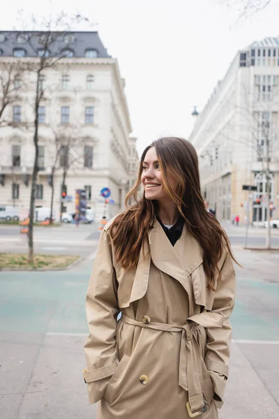 Cheerful young woman in stylish trench coat standing with hands in pockets on street of european city — Stock Photo
