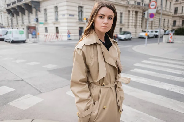 Young woman in stylish trench coat standing with hands in pockets on street of european city — Stock Photo