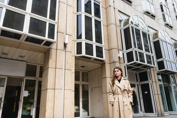 Молода жінка в стильному тренч пальто стоїть руками в кишенях біля будівлі — стокове фото