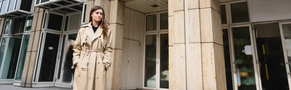 Mujer joven con elegante gabardina de pie con las manos en los bolsillos cerca de la construcción en Viena, pancarta - foto de stock