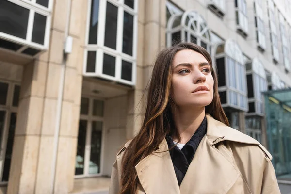 Молода жінка в стильному тренч пальто дивиться далеко біля будівлі — стокове фото