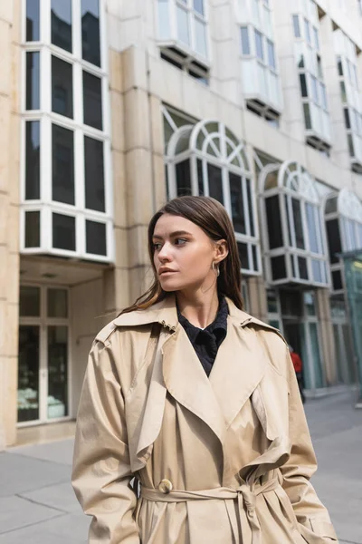 Jeune femme en trench beige regardant loin près du bâtiment — Photo de stock