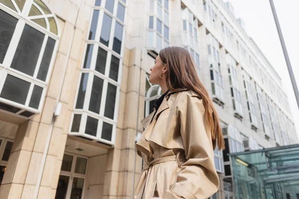 Vista de ángulo bajo de la mujer joven en gabardina de moda mirando el edificio - foto de stock