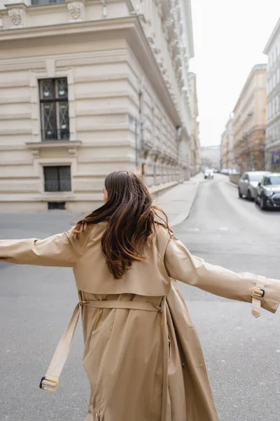 Rückansicht einer Frau im Trenchcoat, die mit ausgestreckten Händen in einer europäischen Stadt spaziert — Stockfoto