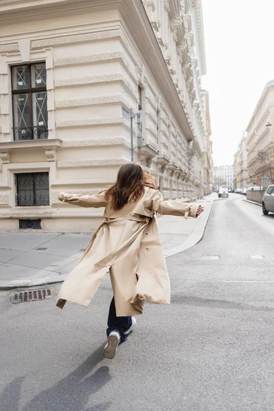 Back view of woman in beige trench coat walking with outstretched hands in european city — Stock Photo