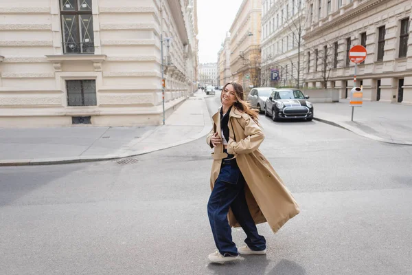 Pleine longueur de gaie jeune femme en trench coat beige marchant sur la rue européenne — Photo de stock