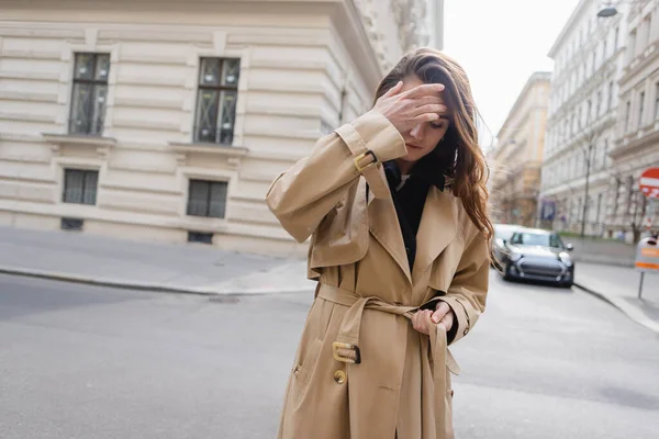 Young woman in beige trench coat adjusting hair on street of european city — Stock Photo