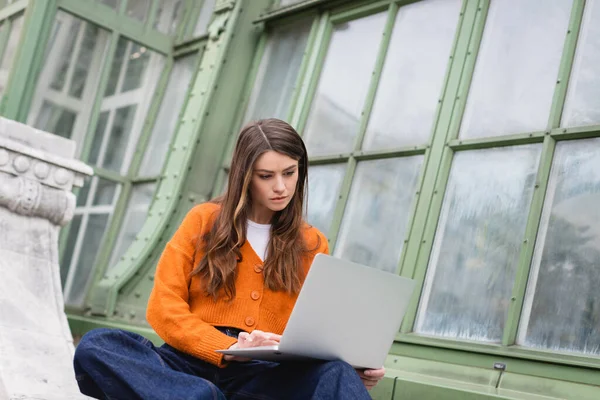 Young freelancer in jeans and orange cardigan using laptop on rooftop of building in vienna — Stock Photo