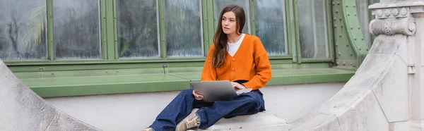 Young woman in jeans and orange cardigan using laptop on rooftop of building, banner — Stock Photo