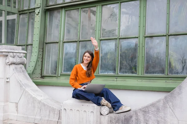 Feliz mujer joven saludando de la mano mientras se utiliza el ordenador portátil en la azotea del edificio en Viena - foto de stock