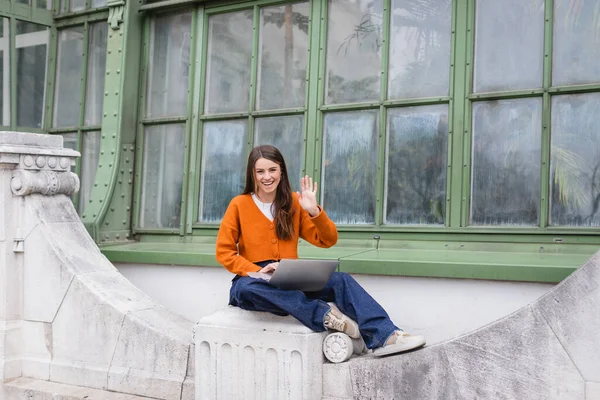 Cheerful young woman waving hand while using laptop on rooftop of building — Stock Photo