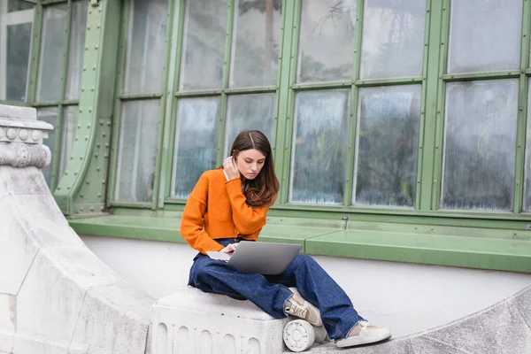 Young woman in jeans and orange cardigan using laptop on rooftop of building — Stock Photo