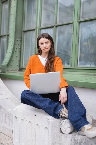 Young woman in jeans and orange cardigan sitting with laptop on rooftop of building — Stock Photo