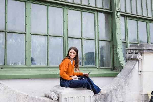 Glückliche junge Freiberuflerin in Jeans und orangefarbener Strickjacke sitzt mit Laptop auf dem Dach des Gebäudes — Stockfoto