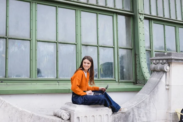 Feliz joven freelancer en jeans y cárdigan naranja sentado con portátil en la azotea del edificio - foto de stock